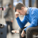 Man at airport sits bending forward with forehead resting on his hand and his suitcase beside him.