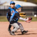 A boy and girl playing with i9 Sports run to catch a baseball, which the boy is about to catch in his glove. A third player can be seen running toward them in the background.