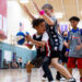 A boy is trying to pass as he's being guarded by a boy from the other team. Other kids on the basketball court are also playing.