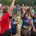 Kids on the sidelines of the field and their coach cheer. The team members are holding trophies.